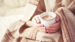  Close-up of hands wrapped in a blanket holding a steaming cup of tea for warmth