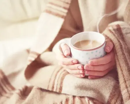  Close-up of hands wrapped in a blanket holding a steaming cup of tea for warmth
