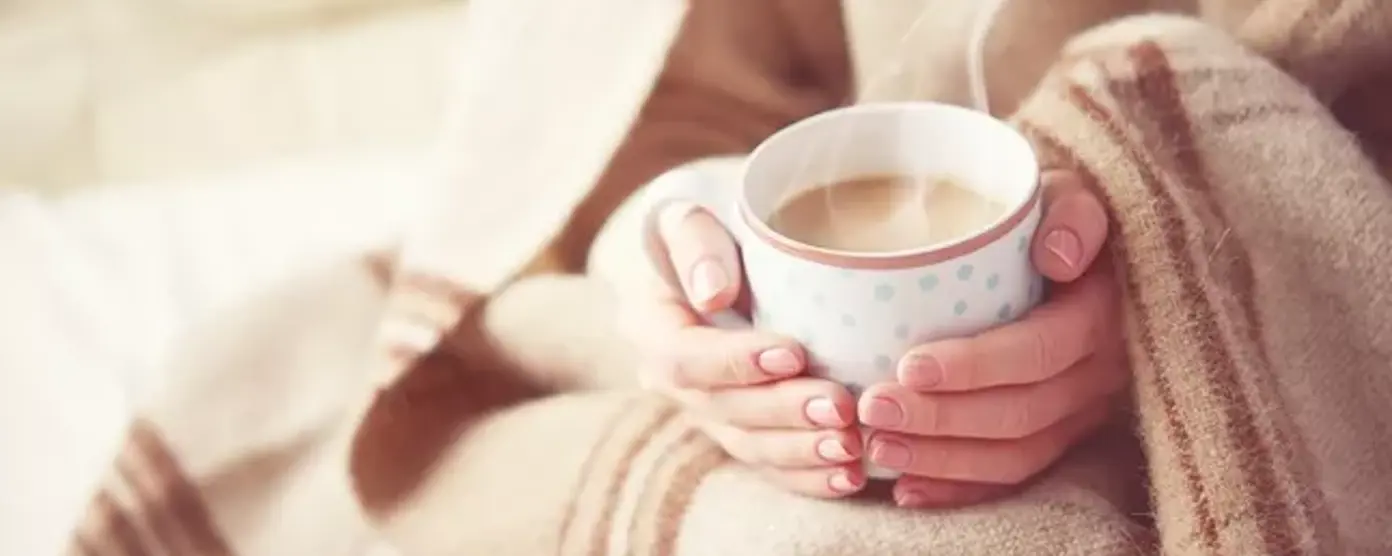  Close-up of hands wrapped in a blanket holding a steaming cup of tea for warmth
