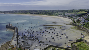 Panoramic aerial view of a turquoise coastal bay with boats, harbor wall, and seaside town on rolling hills
