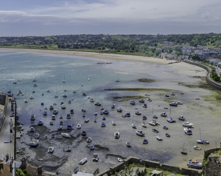 Panoramic aerial view of a turquoise coastal bay with boats, harbor wall, and seaside town on rolling hills