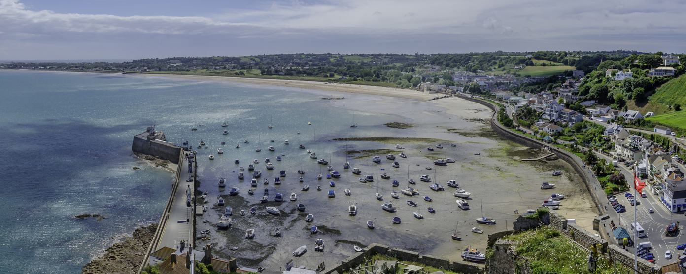 Panoramic aerial view of a turquoise coastal bay with boats, harbor wall, and seaside town on rolling hills