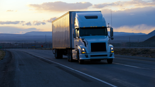 White semi‑truck with box trailer driving on an open highway at dusk with mountains and sunset clouds