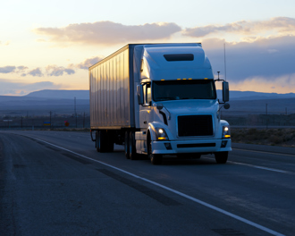 White semi‑truck with box trailer driving on an open highway at dusk with mountains and sunset clouds