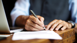 A person signing a document with a pen on a wooden desk beside a laptop