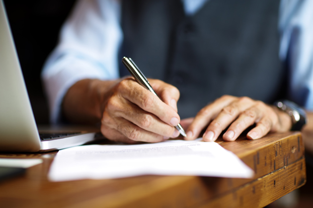 A person signing a document with a pen on a wooden desk beside a laptop