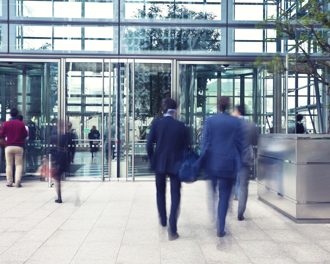 Business professionals actively entering and exiting a modern office building with a sleek glass facade, showcasing corporate life.