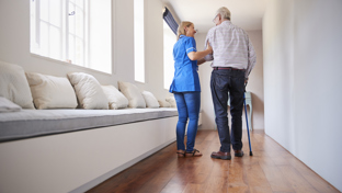 Care worker supporting an elderly man with a cane as they walk down a bright corridor