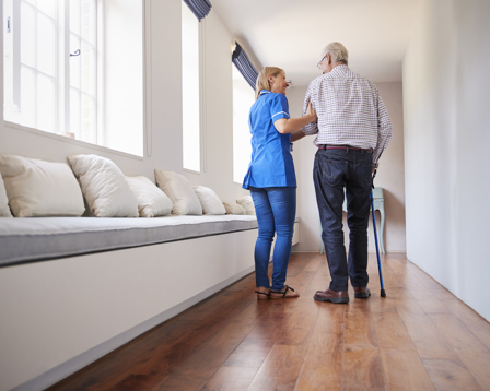 Care worker supporting an elderly man with a cane as they walk down a bright corridor