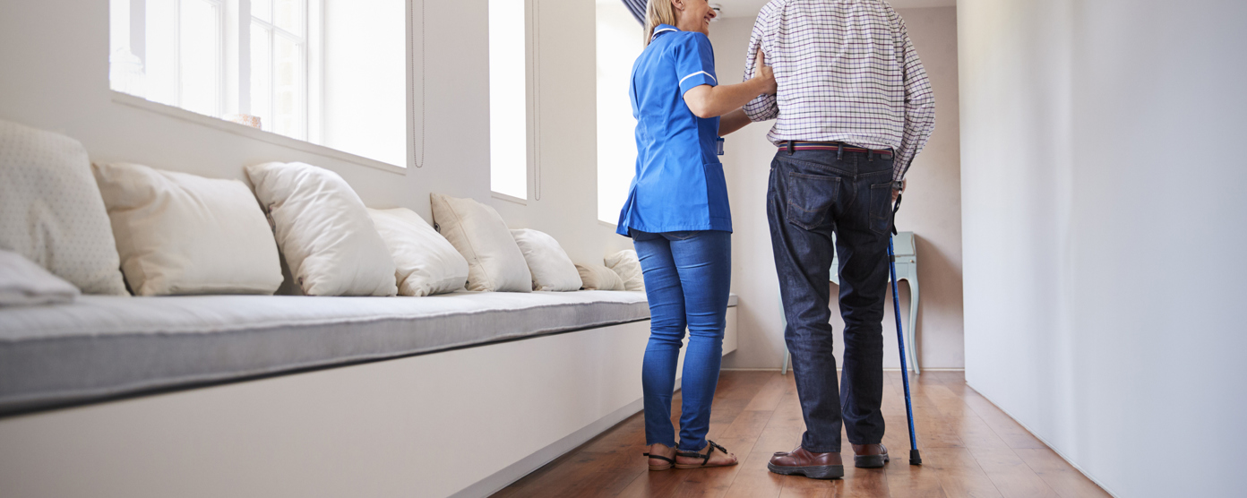 Care worker supporting an elderly man with a cane as they walk down a bright corridor