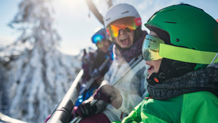 3 people in ski geared on a ski lift with a backdrop of snow laden trees