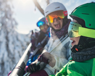 3 people in ski geared on a ski lift with a backdrop of snow laden trees
