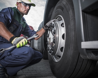 Mechanic in safety gear crouching beside a semi-truck, tightening wheel lug nuts with a large wrench