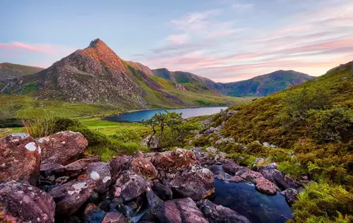 Scenic mountain landscape at sunset with rocky foreground, a calm lake nestled between green hills.