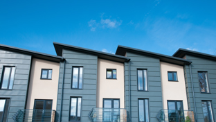 A row of modern townhouses with a clean, contemporary design featuring angled roofs, large vertical windows, and a mix of muted green cladding.