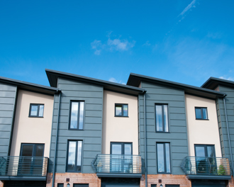 A row of modern townhouses with a clean, contemporary design featuring angled roofs, large vertical windows, and a mix of muted green cladding.