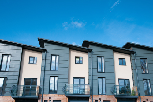 A row of modern townhouses with a clean, contemporary design featuring angled roofs, large vertical windows, and a mix of muted green cladding.