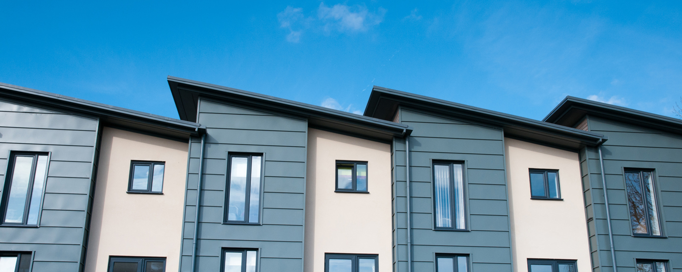 A row of modern townhouses with a clean, contemporary design featuring angled roofs, large vertical windows, and a mix of muted green cladding.