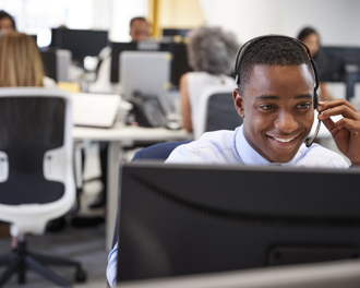 A cheerful young man wearing a headset is seated at a desk in a busy call center or office environment.