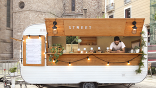 Vintage street-food trailer with wooden serving hatch and lights, vendor leaning at counter in a city square