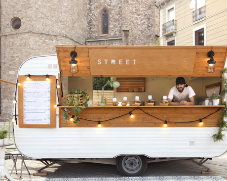 Vintage street-food trailer with wooden serving hatch and lights, vendor leaning at counter in a city square