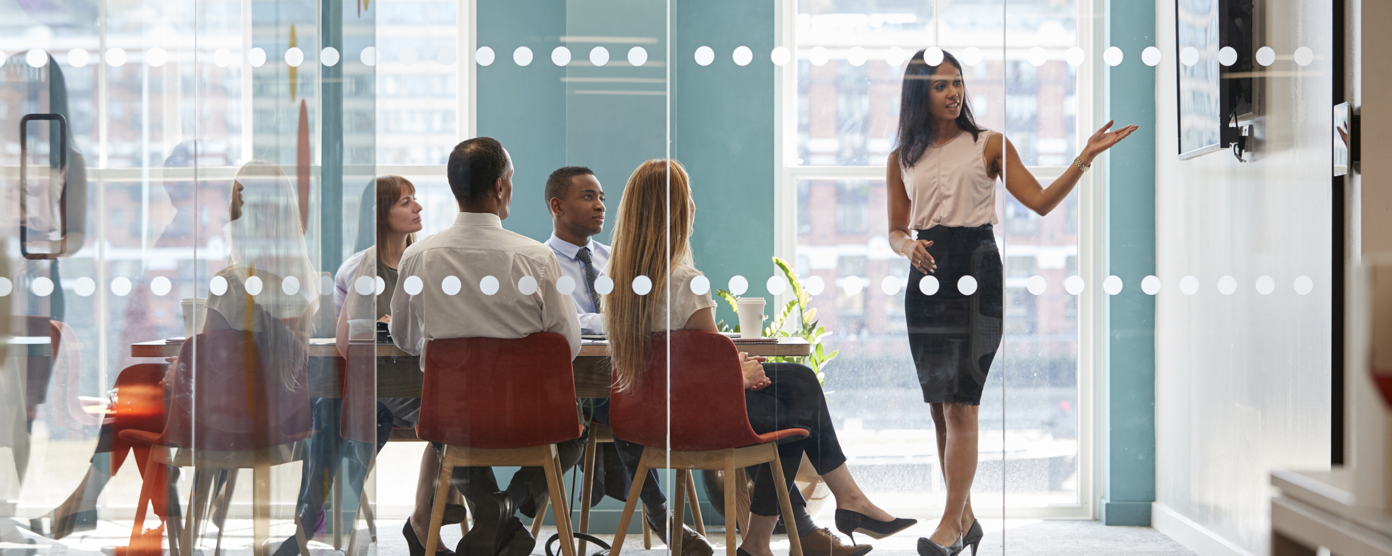 Businesswoman giving a presentation to colleagues in a modern glass-walled conference room during a team meeting.