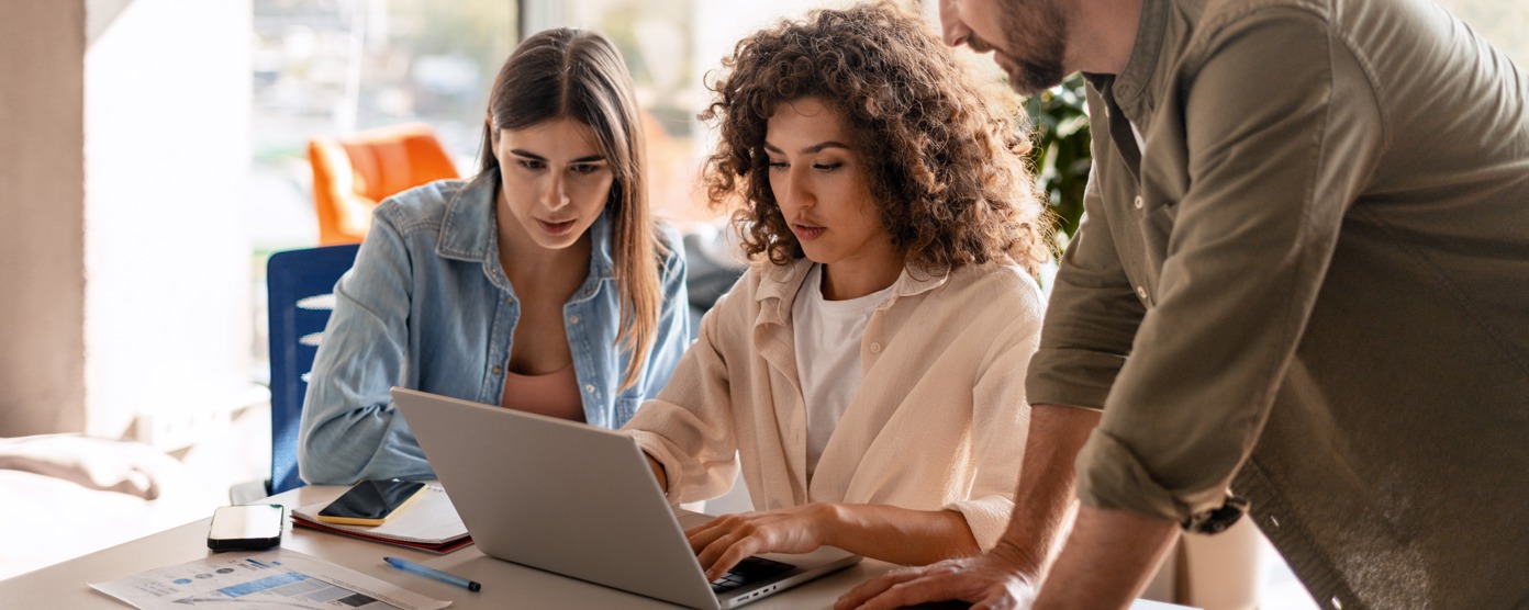 Women typing on a laptop at a desk with 2 others watching