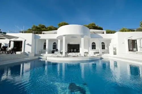 White luxury villa with large swimming pool in the foreground under a clear blue sky.