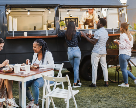 People ordering from a food truck while others eat at outdoor tables in a casual, sunny setting