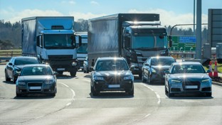 A highway scene showing heavy traffic with cars and large trucks, likely at a junction during peak commuting times.