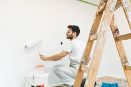 A man wearing white overalls is painting a wall white using a paint roller.
