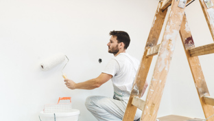 A man wearing white overalls is painting a wall white using a paint roller.