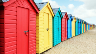 A row of brightly painted beach huts stands on sandy ground, each in different pastel and vibrant colors.