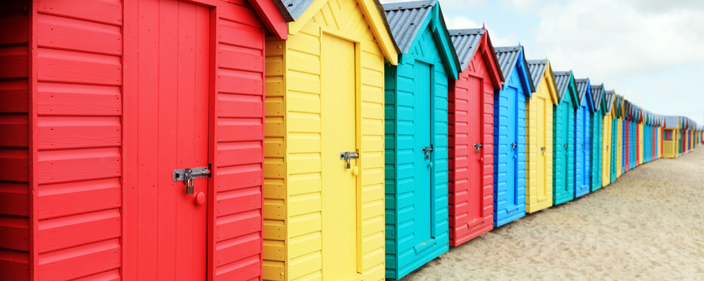 A row of brightly painted beach huts stands on sandy ground, each in different pastel and vibrant colors.
