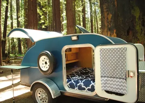 Teardrop camper with open hatch showing renovated interior and bed at a forest campsite