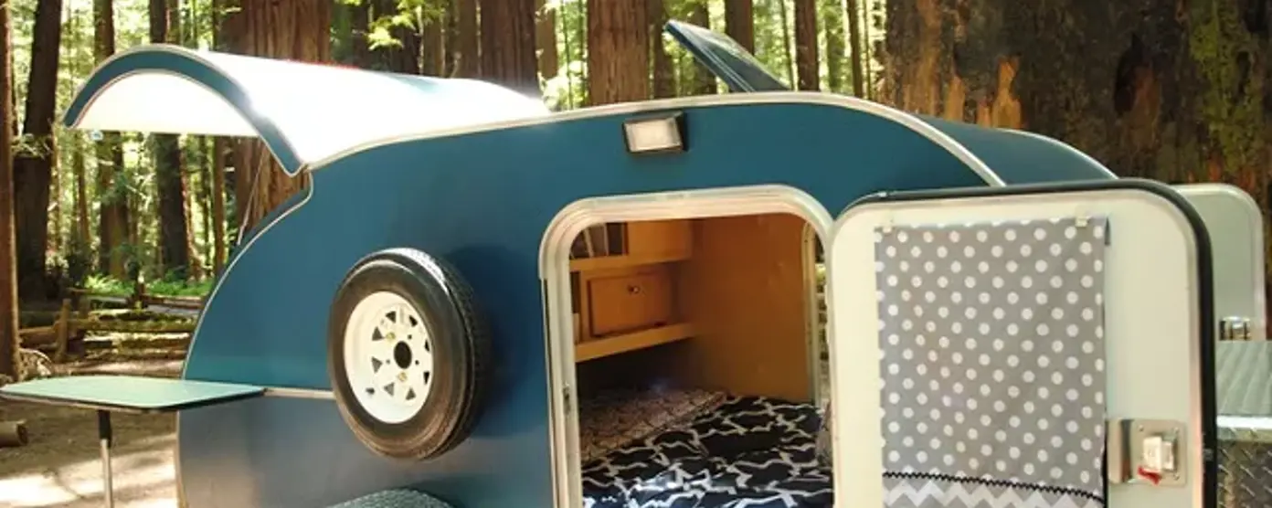 Teardrop camper with open hatch showing renovated interior and bed at a forest campsite