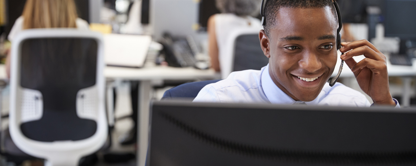 A cheerful customer service agent wearing a headset engages with clients at a computer in a lively office environment.