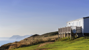 Static caravan with wooden decking on a grassy coastal cliff, overlooking the sea under a clear blue sky.