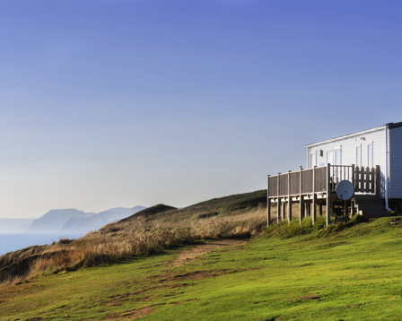 Static caravan with wooden decking on a grassy coastal cliff, overlooking the sea under a clear blue sky.