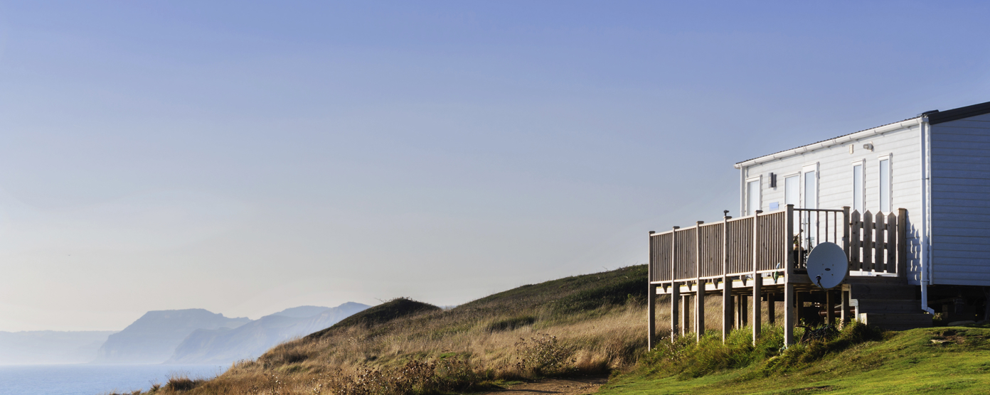 Static caravan with wooden decking on a grassy coastal cliff, overlooking the sea under a clear blue sky.