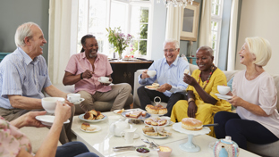  Group of smiling seniors sharing afternoon tea and cakes in a bright living room, sitting around a coffee table and chatting together