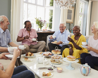  Group of smiling seniors sharing afternoon tea and cakes in a bright living room, sitting around a coffee table and chatting together