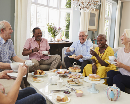  Group of smiling seniors sharing afternoon tea and cakes in a bright living room, sitting around a coffee table and chatting together