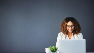 A smiling woman with curly hair and glasses works on a laptop at a desk adorned with a plant and a coffee mug.