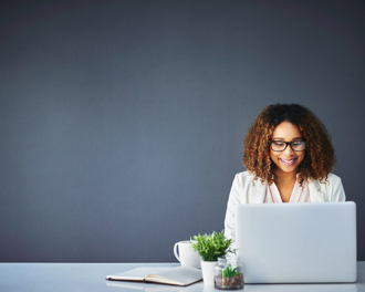 A smiling woman with curly hair and glasses works on a laptop at a desk adorned with a plant and a coffee mug.