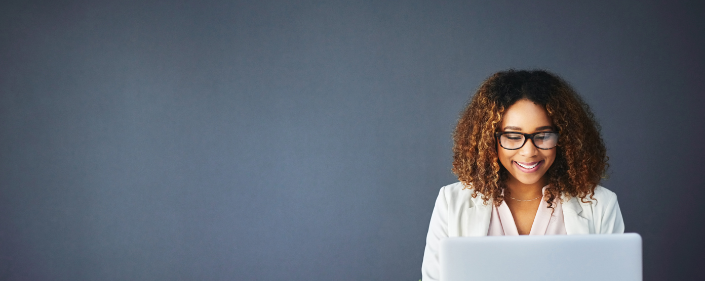 A smiling woman with curly hair and glasses works on a laptop at a desk adorned with a plant and a coffee mug.