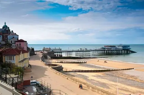 View of Cromer’s sandy beach and Victorian pier on the Norfolk coast under a bright, partly cloudy sky.
