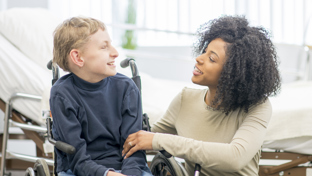Caring woman in a wheelchair chatting warmly with a smiling boy in a wheelchair in a bright room, offering support and encouragement