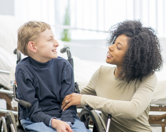 Caring woman in a wheelchair chatting warmly with a smiling boy in a wheelchair in a bright room, offering support and encouragement