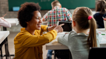 Smiling teacher kneeling beside a student’s desk, pointing at a tablet and offering guidance in a bright classroom with a chalkboard in the background